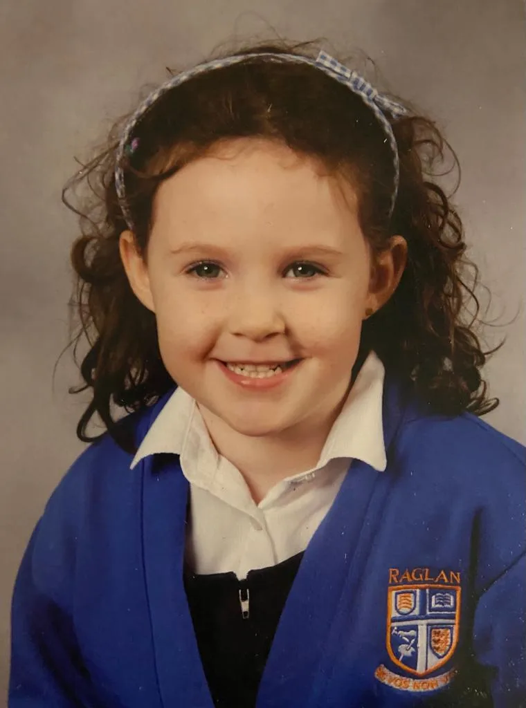 A young girl with curly hair and a hairband smiling in a school portrait, wearing a blue jumper with a Raglan school crest